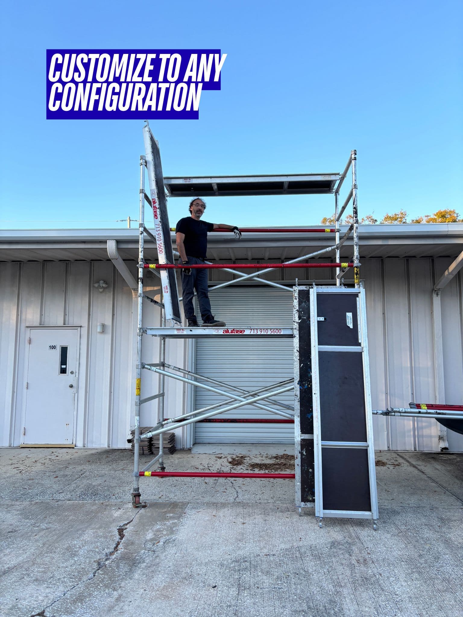 Man standing on a customizable aluminum scaffolding tower in front of a white industrial building.
