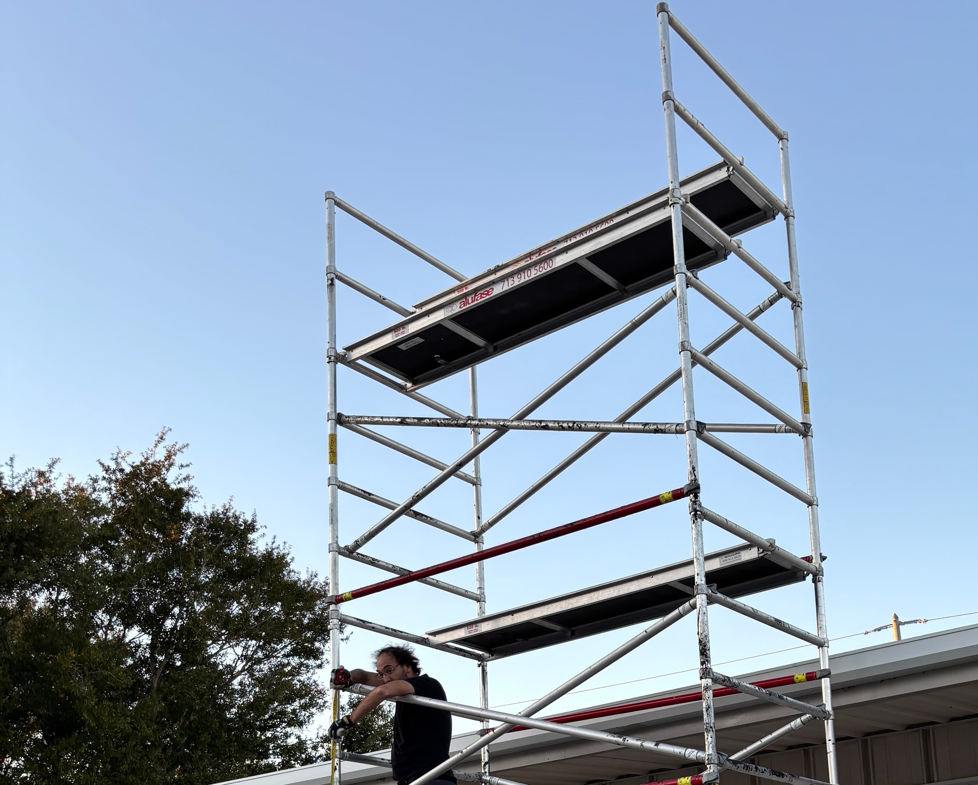 Man in black shirt works on a tall silver scaffolding tower against a blue sky.