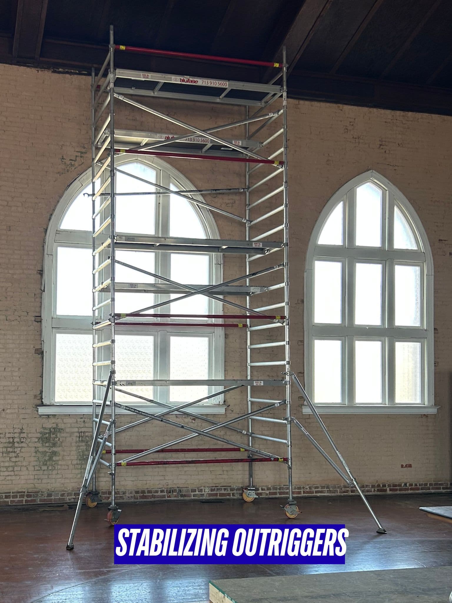 Tall aluminum scaffolding tower with stabilizing outriggers stands in a room with arched windows.