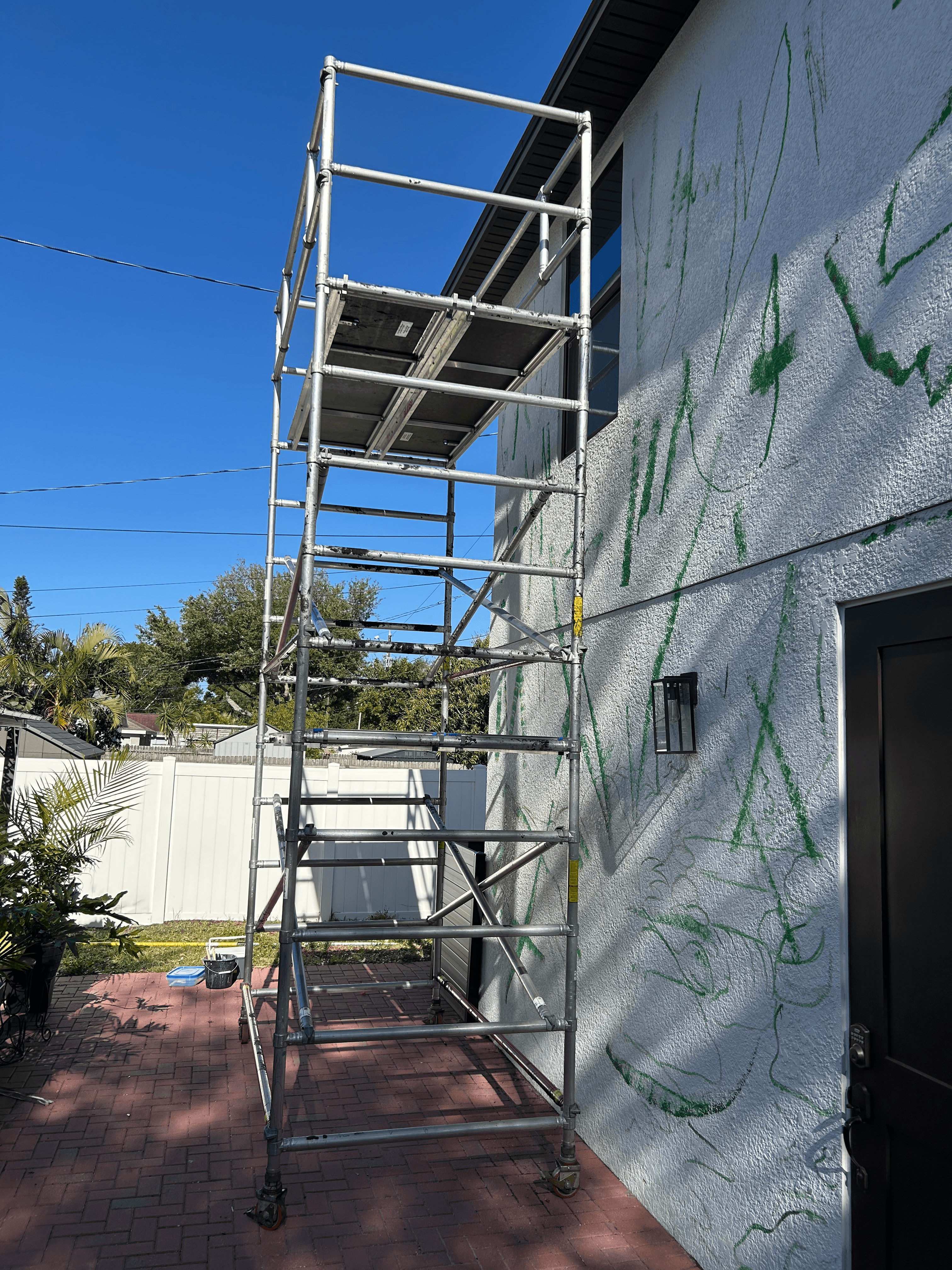 Metal scaffolding stands against a white textured wall covered in green graffiti under a blue sky.