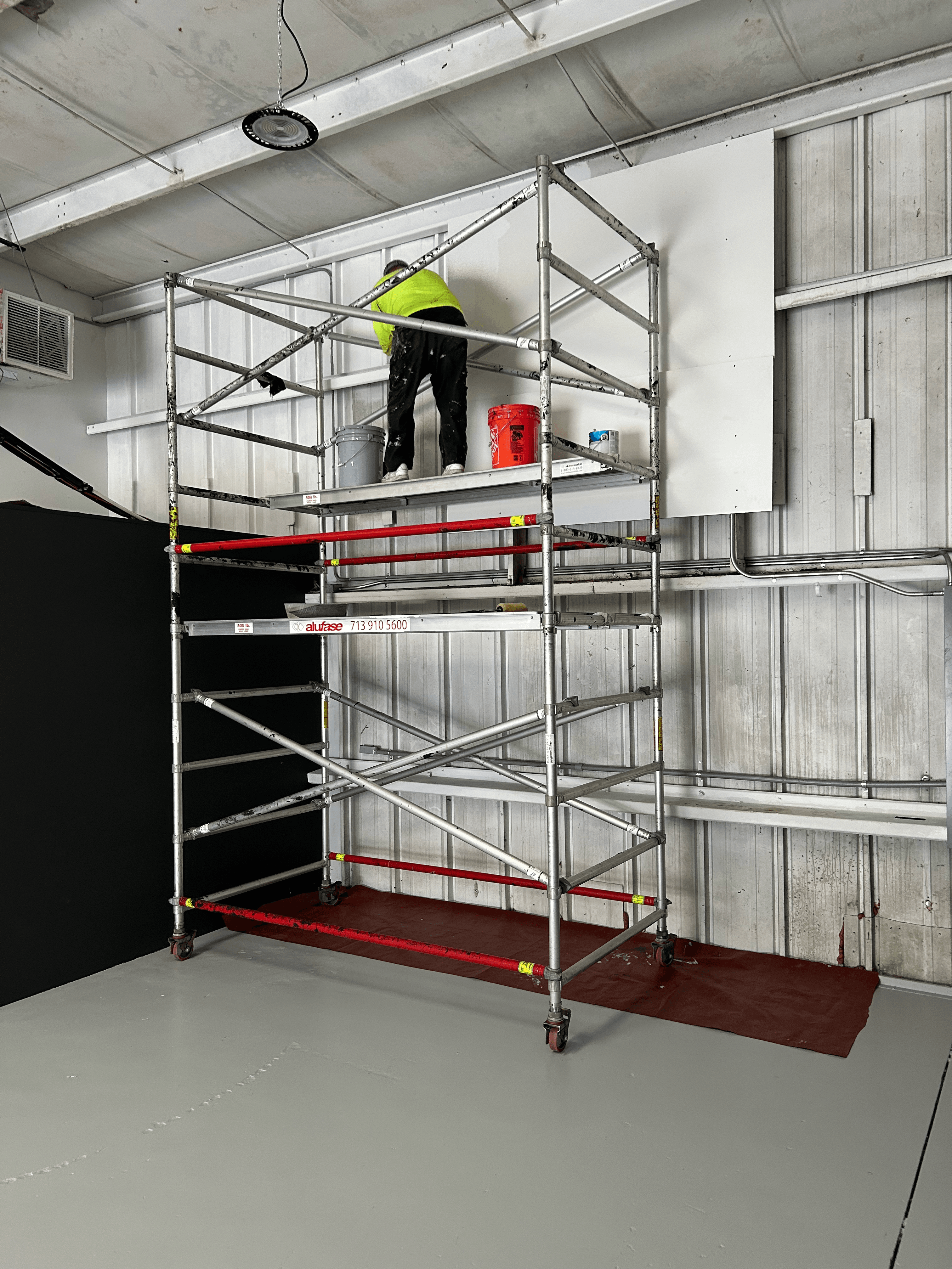 Worker in high-visibility gear stands on a tall metal scaffolding tower inside a large building.