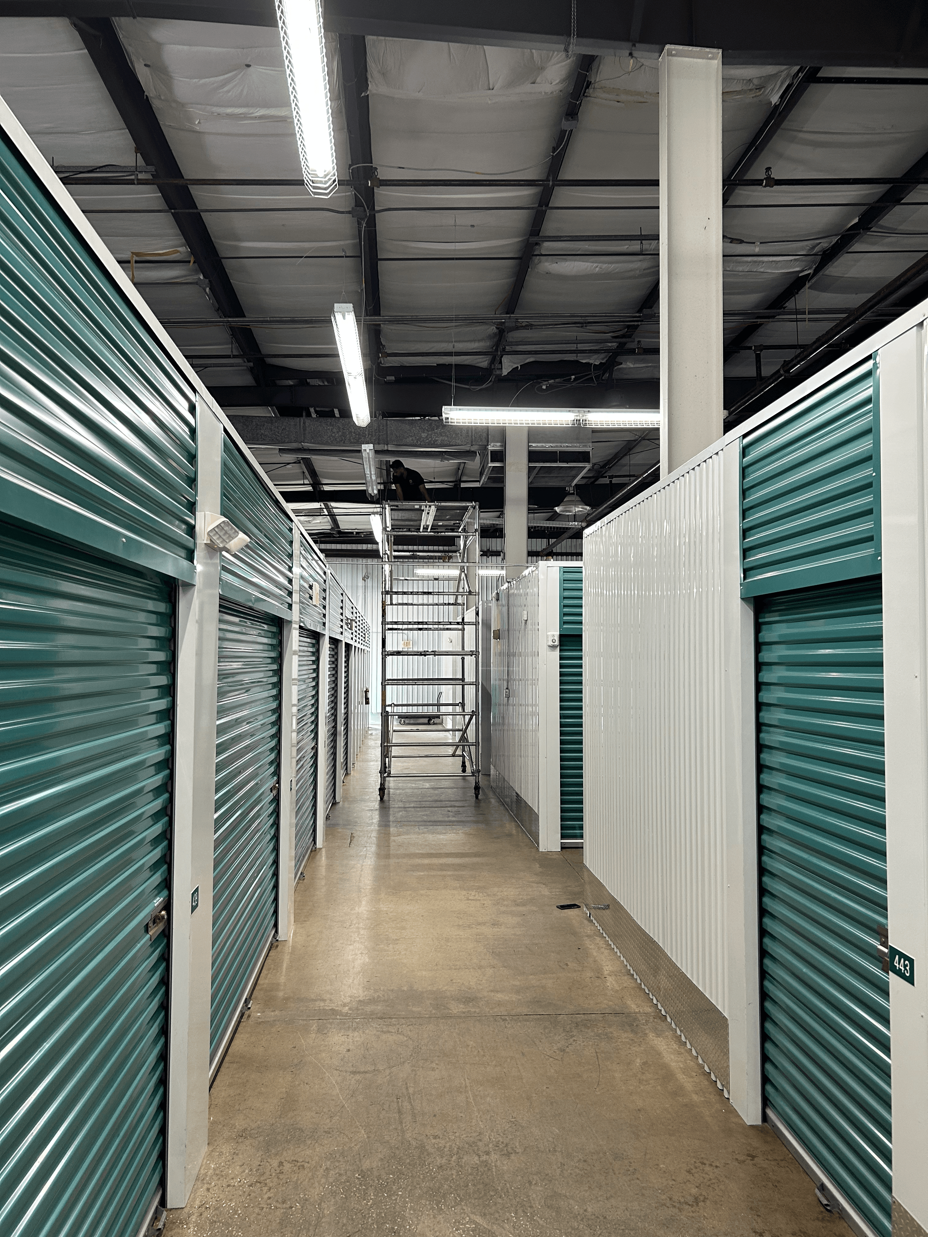 Long storage facility hallway with teal corrugated metal doors and bright industrial overhead lighting.